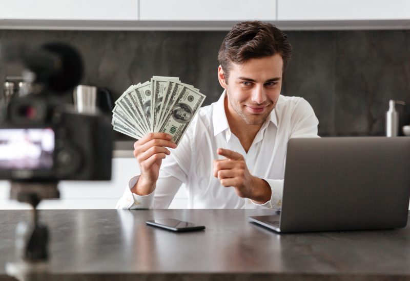 Smiling young man filming his video blog episode about new tech devices while sitting at the kitchen table with laptop and showing bunch of money banknotes