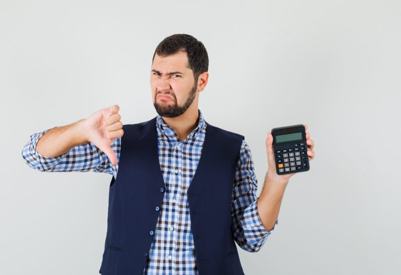 Young man holding calculator, showing thumb down in shirt, vest and looking dissatisfied , front view.